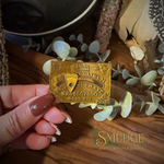 A hand with manicured nails holds a small gold-colored novelty card or charm featuring vintage-style text and a map pin design, set against a rustic wooden background with decorative eucalyptus leaves and dried botanicals. The brand name 'SMUDGE' is visible in the bottom right corner.