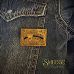 A rectangular bronze-toned metal plaque featuring occult-style text and symbols, displayed on a dark blue denim jacket. The plaque is positioned over the pocket area of the denim.