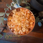 A top-down view of a wooden bowl filled with translucent orange and amber crystal salt, styled on a dark wooden surface with green eucalyptus leaves. The brand name 'SMUDGE' is visible in the bottom-left corner.