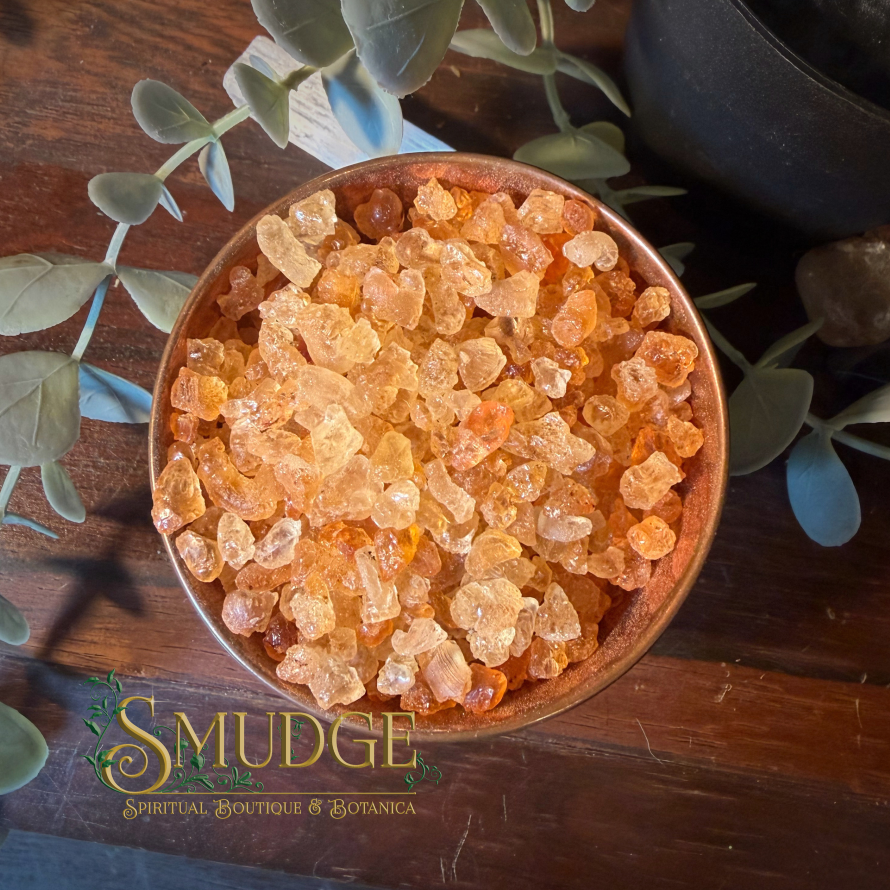 A top-down view of a wooden bowl filled with translucent orange and amber crystal salt, styled on a dark wooden surface with green eucalyptus leaves. The brand name 'SMUDGE' is visible in the bottom-left corner.