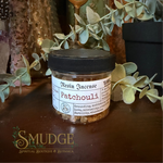 A small clear jar of resin incense labeled 'Patchouli' with a black lid, placed on a dark wooden surface with decorative eucalyptus leaves and rustic decor in the background. The Smudge Spiritual Boutique & Botanica logo appears at the bottom left.
