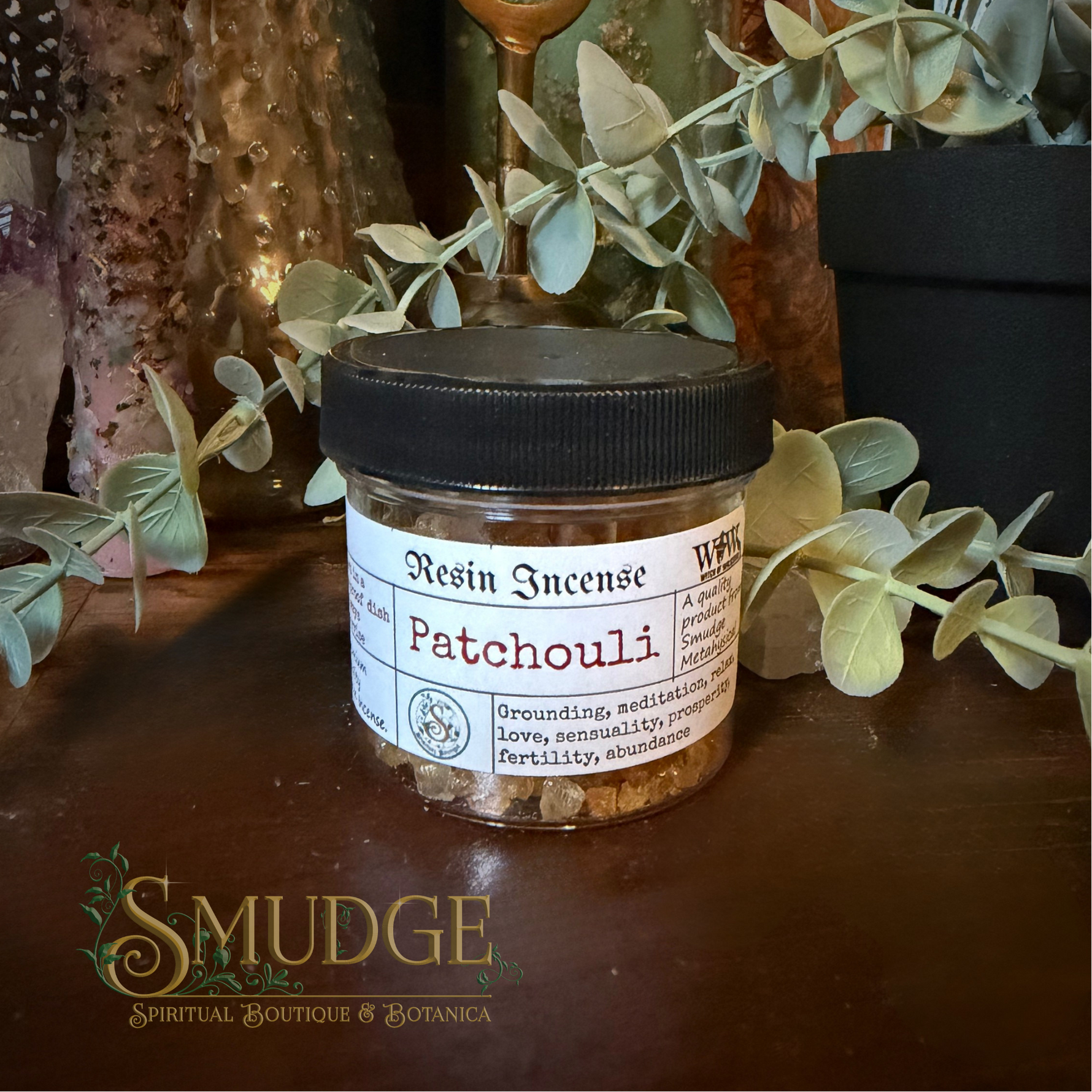 A small clear jar of resin incense labeled 'Patchouli' with a black lid, placed on a dark wooden surface with decorative eucalyptus leaves and rustic decor in the background. The Smudge Spiritual Boutique & Botanica logo appears at the bottom left.