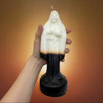 A hand with a tattooed forearm holds a religious candle shaped like the Virgin Mary. The candle is white on the upper half with a black lower half and a thin wick at the top, shown against a plain white background.