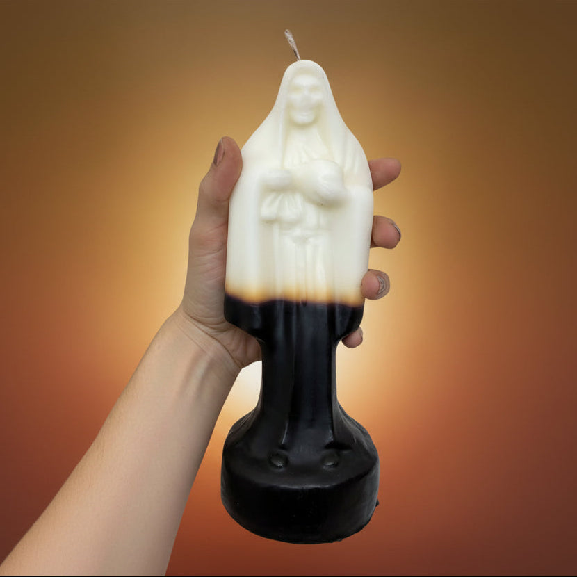 A hand with a tattooed forearm holds a religious candle shaped like the Virgin Mary. The candle is white on the upper half with a black lower half and a thin wick at the top, shown against a plain white background.