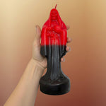 A hand with visible tattoos holds a red and black gradient candle shaped like a robed figure, shown against a plain white background.
