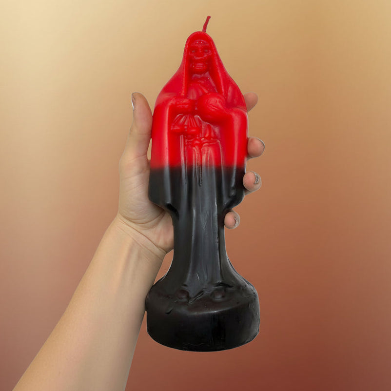 A hand with visible tattoos holds a red and black gradient candle shaped like a robed figure, shown against a plain white background.