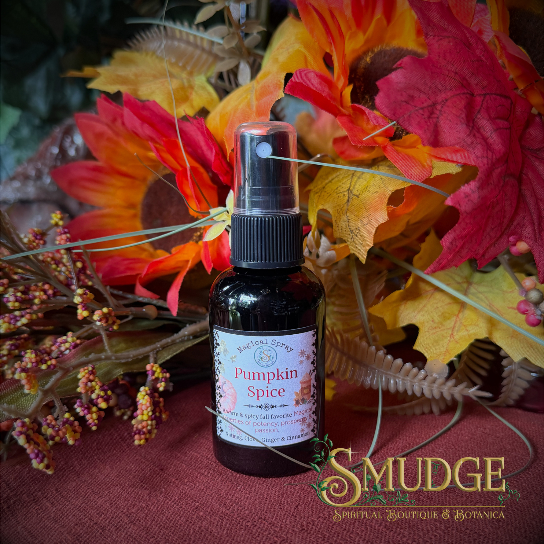 A bottle of Pumpkin Spice essential oil or room spray by Smudge Spiritual Boutique & Botanica, styled with autumn leaves and sunflowers on a red fabric surface.