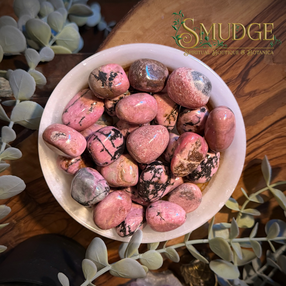 A bowl filled with polished pink and brown Rhodonite stones, photographed from above on a wooden surface with decorative greenery. The brand name 'SMUDGE' is visible in the upper right.