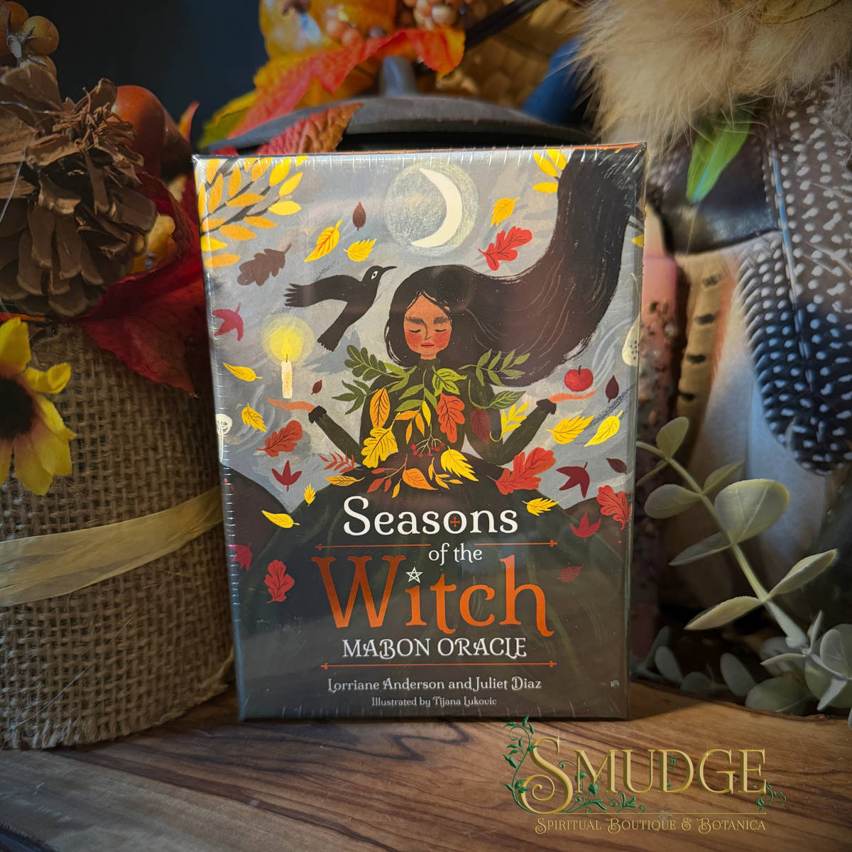 The 'Seasons of the Witch' Marion Oracle card deck box is displayed on a wooden surface surrounded by autumn-themed decorative props, including a burlap pot with a sunflower, dried leaves, and feathers. The box features an illustration of a witch with a cauldron, surrounded by falling autumn leaves under a crescent moon.