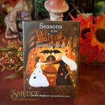 Seasons of the Witch: Autumn Tarot card deck box displayed on a red fabric surface with autumn-themed decorative props including sunflowers, dried flowers, and a mushroom figurine in the background.