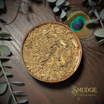 Dry herbs in a bowl on a wooden surface with eucalyptus leaves and a peacock feather, featuring the brand 'Smudge'.