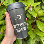 A hand holds a black reusable travel tumbler with a white crescent moon and stars graphic and the words 'WITCHES BREW' in white, set against a background of green leaves.