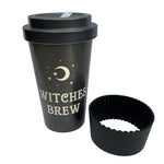 A black reusable travel tumbler with a matching black lid, printed with a gold crescent moon, stars, and the words 'WITCHES BREW,' shown next to a black silicone cupcake liner on a white background.