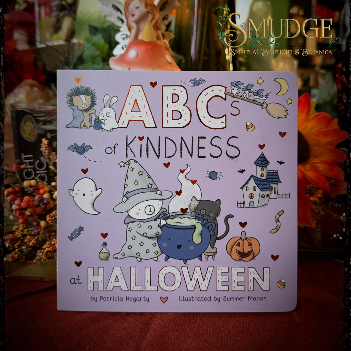 The front cover of the children's book 'ABCs of Kindness at Halloween' by Patricia Hegarty and Summer Macon, featuring a light purple background with whimsical Halloween illustrations including a witch, ghost, cat, and haunted house. The book is displayed on a dark red surface with a blurred background of autumn decorations.