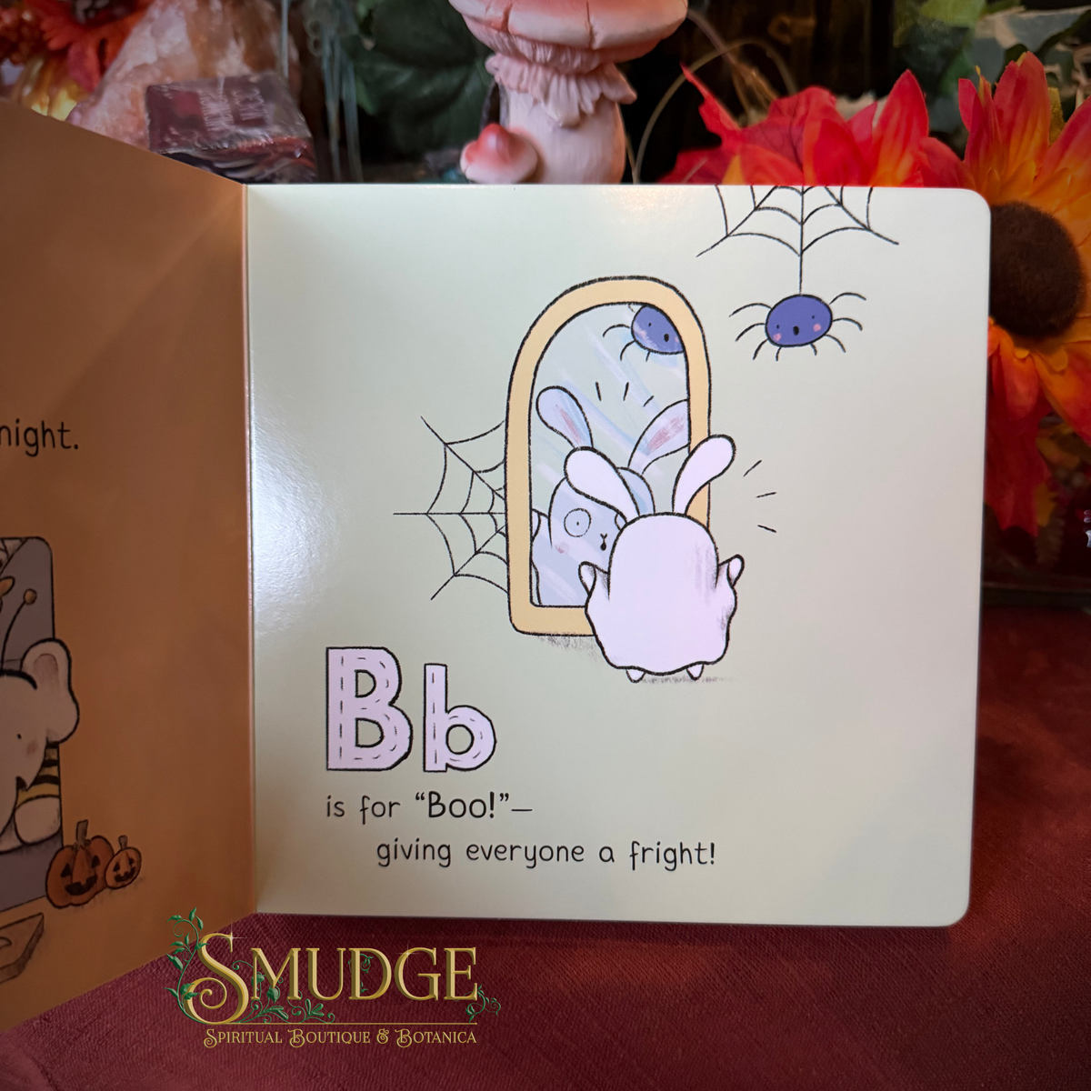 An open children's board book showing a Halloween-themed page with a white bunny looking into a mirror, spiderwebs, a spider on a cobweb, and the text 'Bb is for "Boo!" giving everyone a fright!'. The book is photographed on a dark red surface with flowers and figurines in the background.