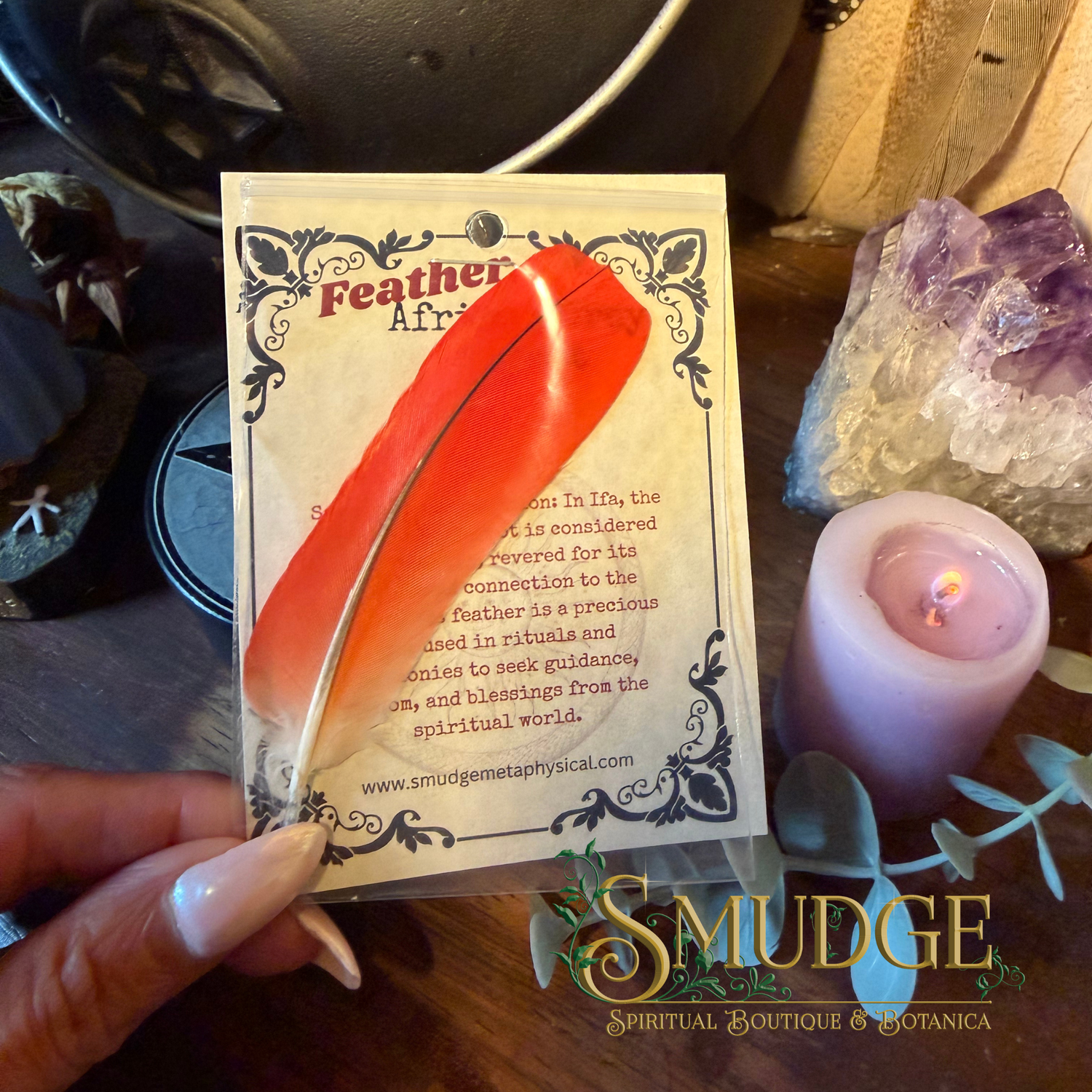 A hand holds a card featuring a single bright orange-red feather with a printed description card behind it. The scene includes a lit pink candle, crystals, and a dark wooden surface, with a visible Smudge branding watermark at the bottom.