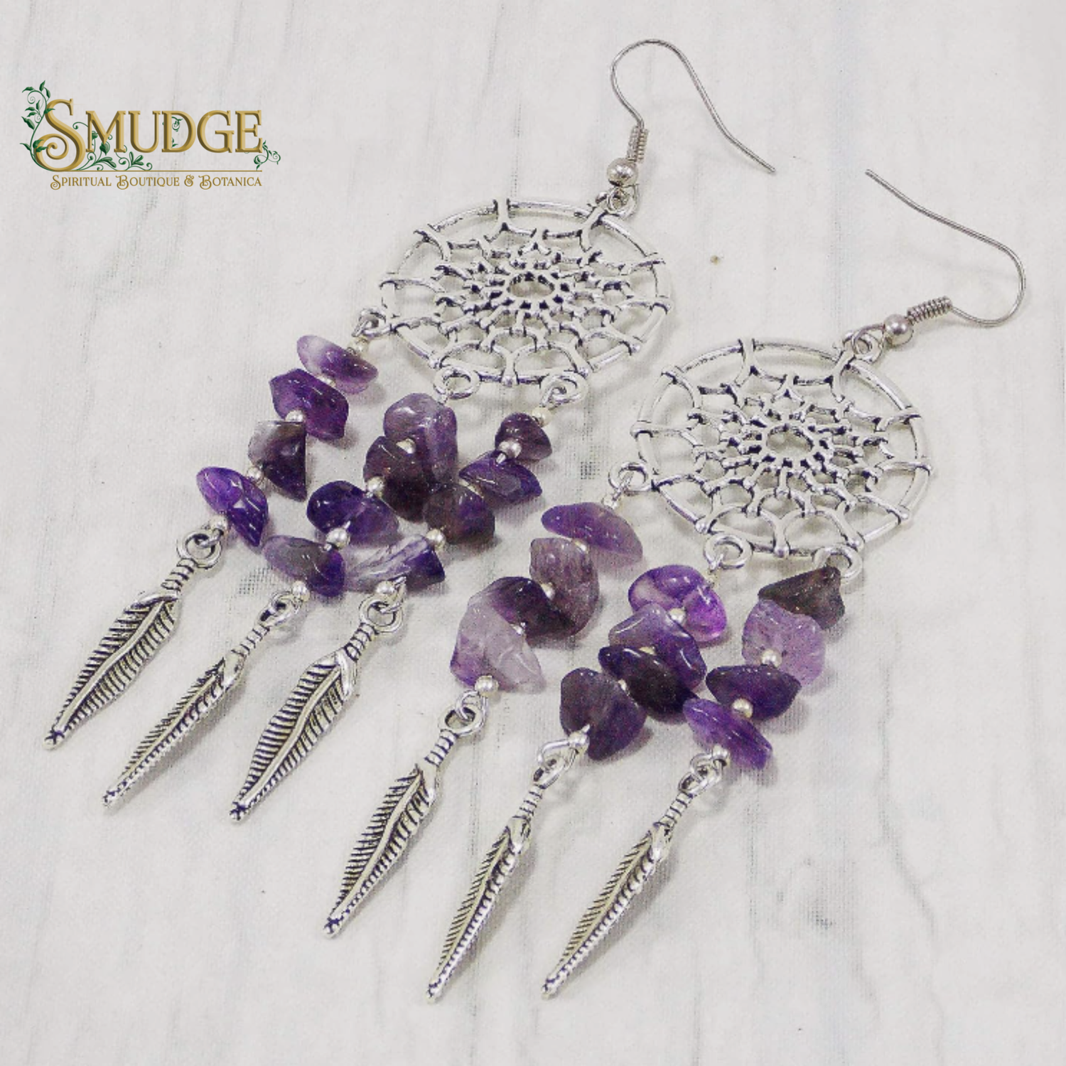 A pair of dreamcatcher-inspired dangle earrings with silver-toned metal web motifs, purple amethyst chip beads, and silver feather charms, displayed on a light marble surface. The Smudge Spiritual Boutique & Botanica logo is visible in the top left corner.