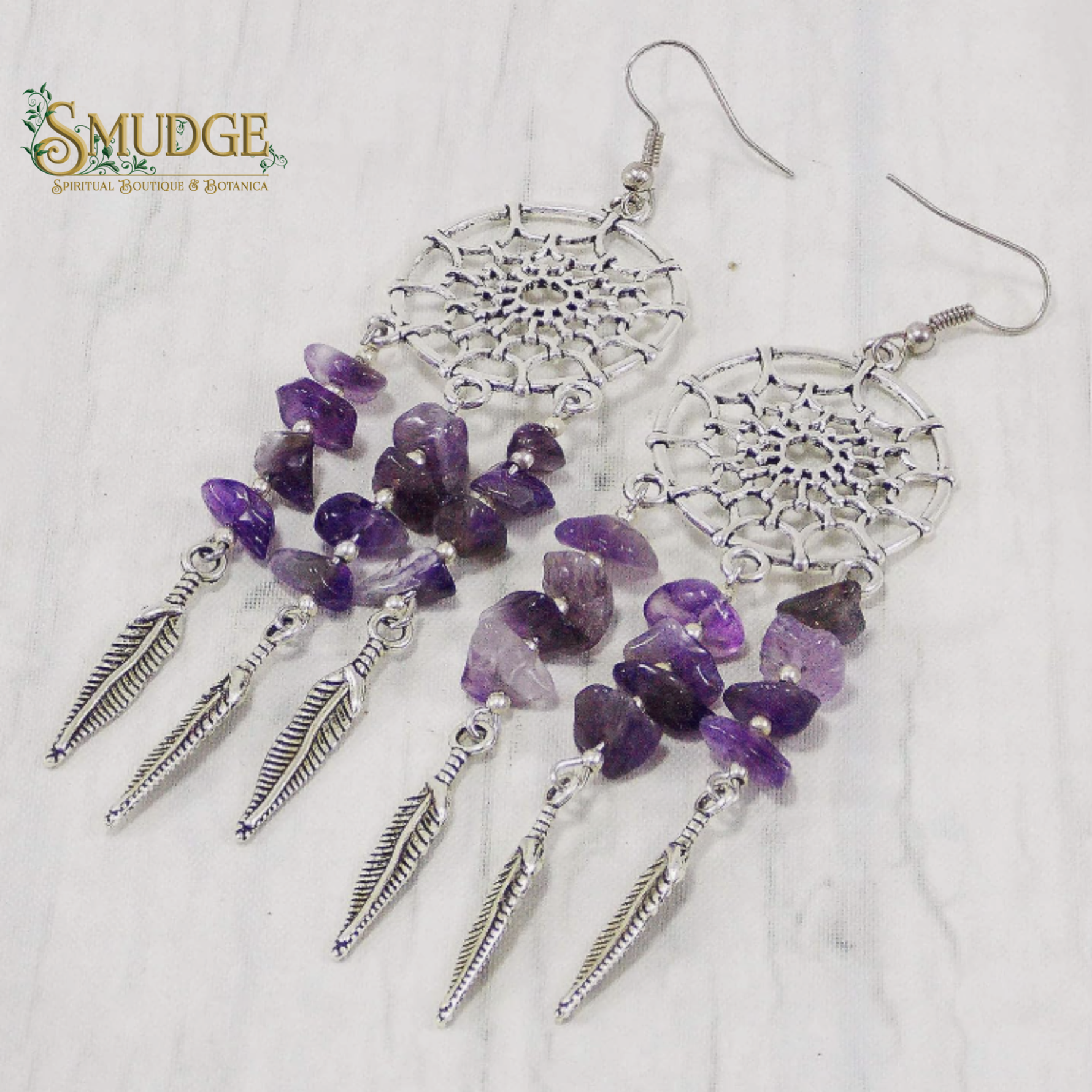 A pair of dreamcatcher-inspired dangle earrings with silver-toned metal web motifs, purple amethyst chip beads, and silver feather charms, displayed on a light marble surface. The Smudge Spiritual Boutique & Botanica logo is visible in the top left corner.