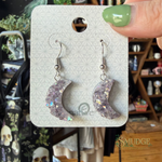 A pair of crescent moon drop earrings with glittery lavender-purple resin and iridescent flecks, displayed on a white backing card held by a hand with green nail polish. The background is a blurry shop interior.