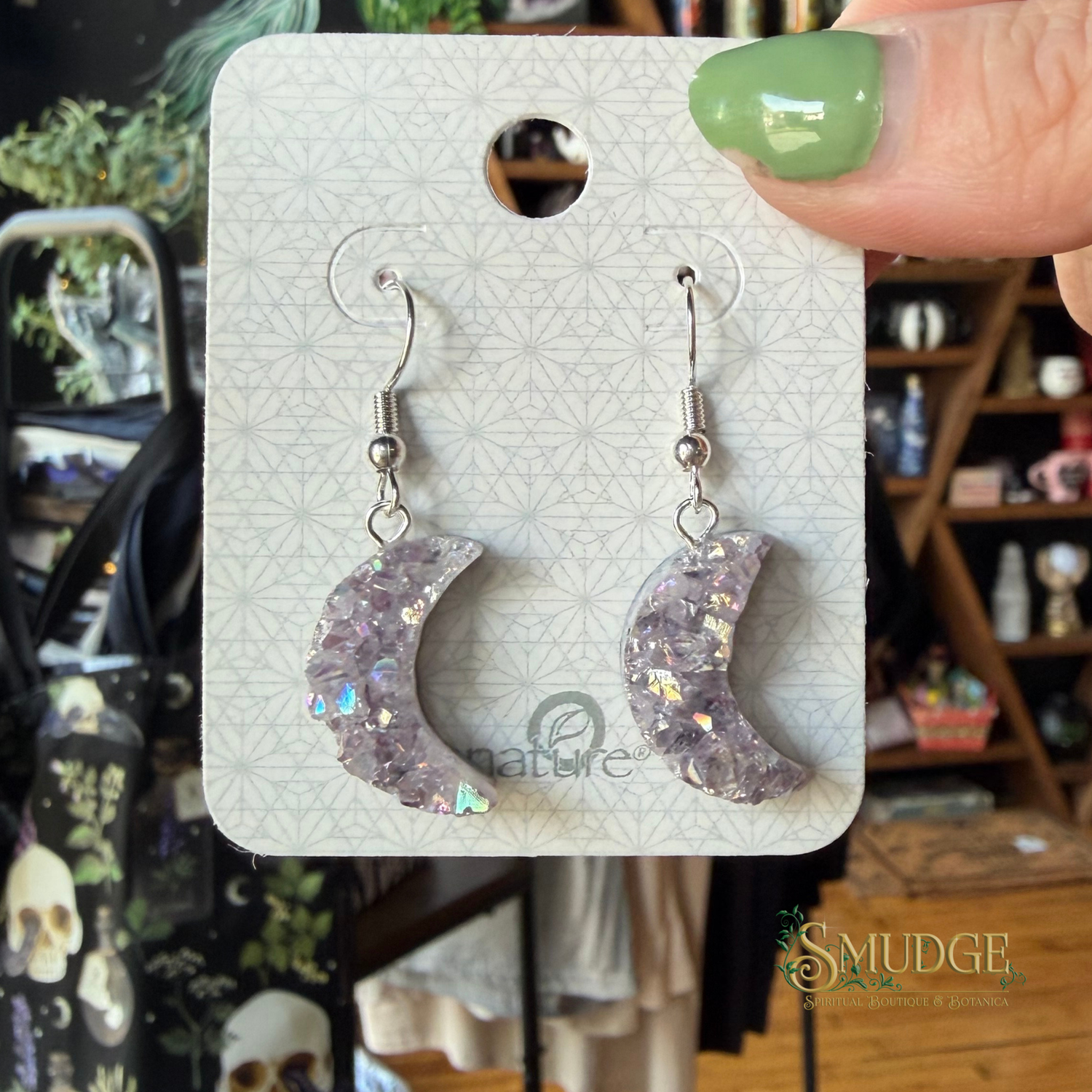 A pair of crescent moon drop earrings with glittery lavender-purple resin and iridescent flecks, displayed on a white backing card held by a hand with green nail polish. The background is a blurry shop interior.