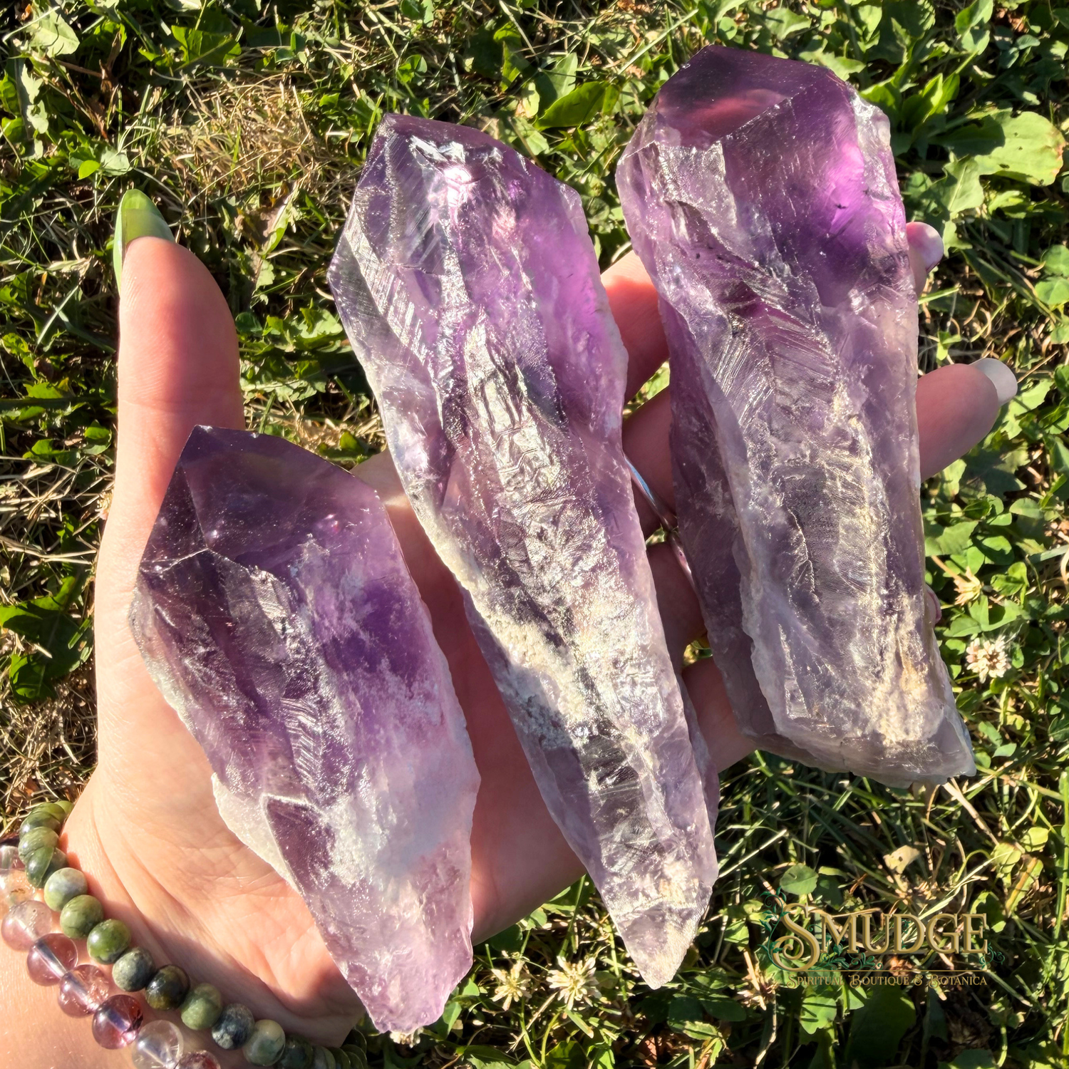Three raw amethyst crystal points held in a person's hand outdoors on grass, with a bracelet visible on the wrist.