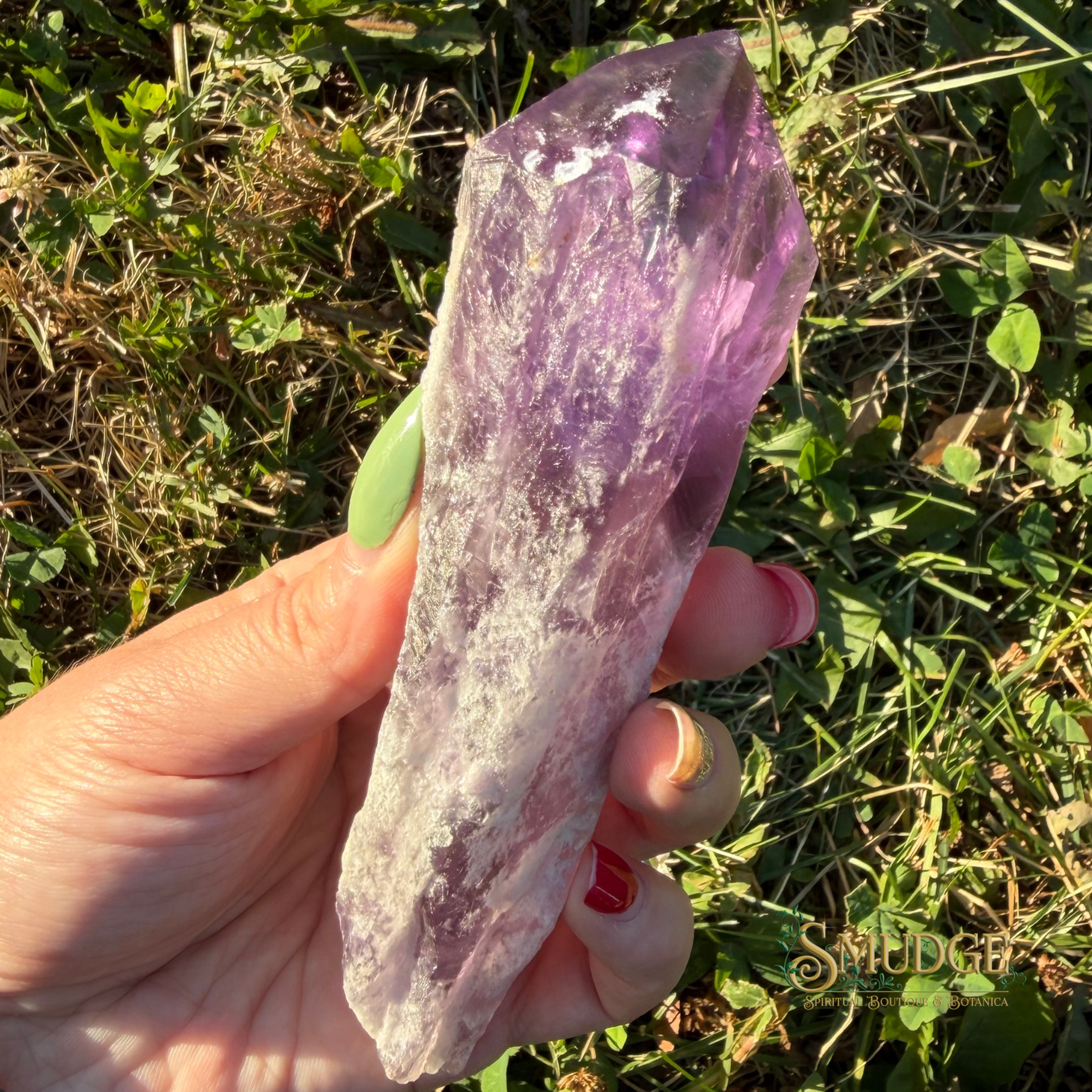A hand holding a raw purple amethyst crystal point outdoors against a background of green grass and foliage.