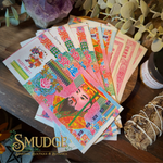 A fan of colorful Chinese New Year paper money or banknote-style decorative paper pieces spread on a wooden surface, with a smudge bundle, greenery, and a jar nearby. The Smudge Spiritual Boutique & Botanica logo appears at the bottom left.