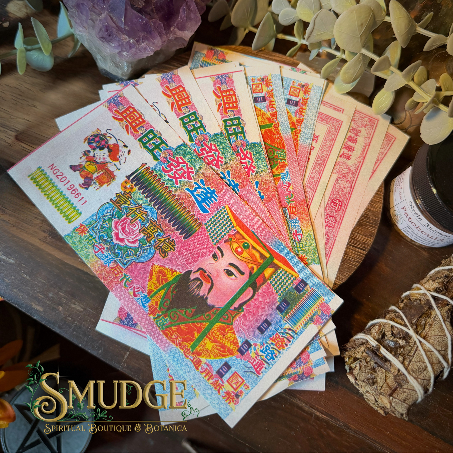 A fan of colorful Chinese New Year paper money or banknote-style decorative paper pieces spread on a wooden surface, with a smudge bundle, greenery, and a jar nearby. The Smudge Spiritual Boutique & Botanica logo appears at the bottom left.