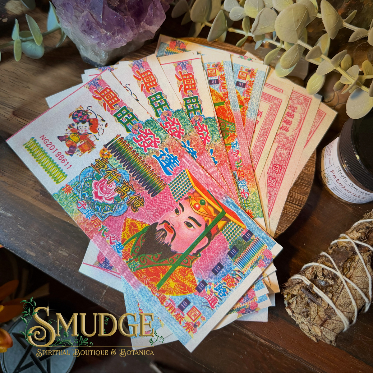 A fan of colorful Chinese New Year paper money or banknote-style decorative paper pieces spread on a wooden surface, with a smudge bundle, greenery, and a jar nearby. The Smudge Spiritual Boutique & Botanica logo appears at the bottom left.