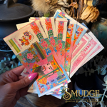 A hand with pink nail polish holds a fan of colorful Chinese New Year paper money or lucky charms featuring traditional illustrations and red Chinese characters. The background is a rustic, decorated setting with dried flowers and plants, and a 'SMUDGE Spiritual Goods & Services' logo is visible in the bottom right corner.