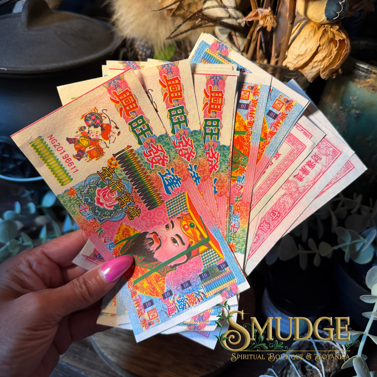A hand with pink nail polish holds a fan of colorful Chinese New Year paper money or lucky charms featuring traditional illustrations and red Chinese characters. The background is a rustic, decorated setting with dried flowers and plants, and a 'SMUDGE Spiritual Goods & Services' logo is visible in the bottom right corner.