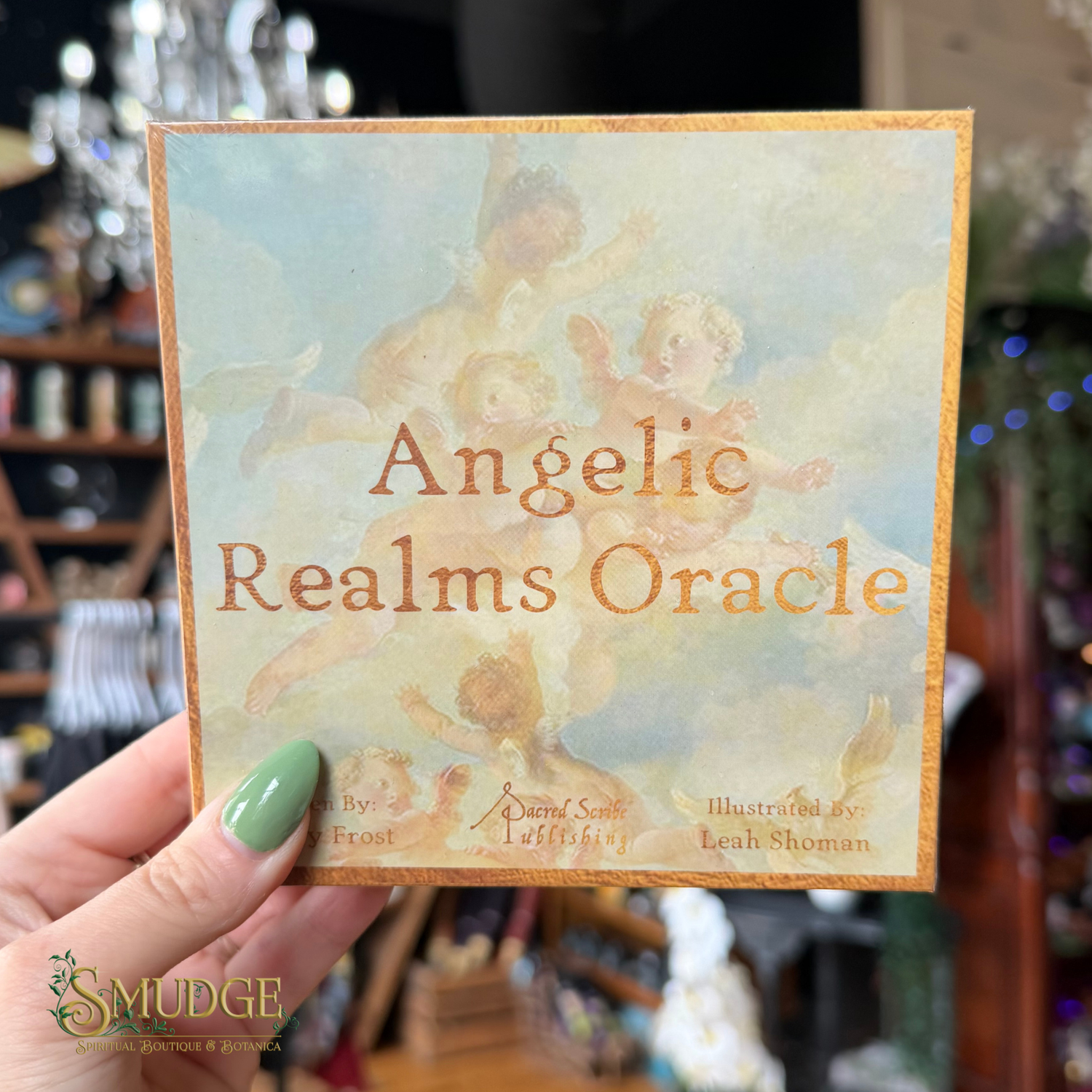A hand with green nail polish holds a square card or box cover for the 'Angelic Realms Oracle' by Digital Sentie, featuring a classical painting of cherubs with a gold border. The background is a blurry shop interior.
