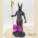 An Egyptian Anubis figurine with a dark gray fur coat, holding a gold-colored scythe, standing on a textured black base with a pink flower accent. The figurine is displayed on a light-colored stone slab against a neutral background.