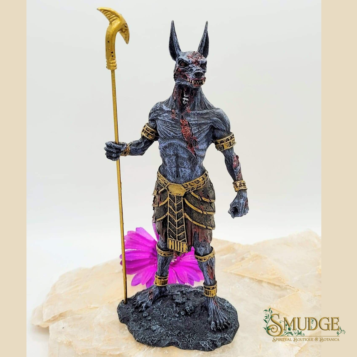 An Egyptian Anubis figurine with a dark gray fur coat, holding a gold-colored scythe, standing on a textured black base with a pink flower accent. The figurine is displayed on a light-colored stone slab against a neutral background.