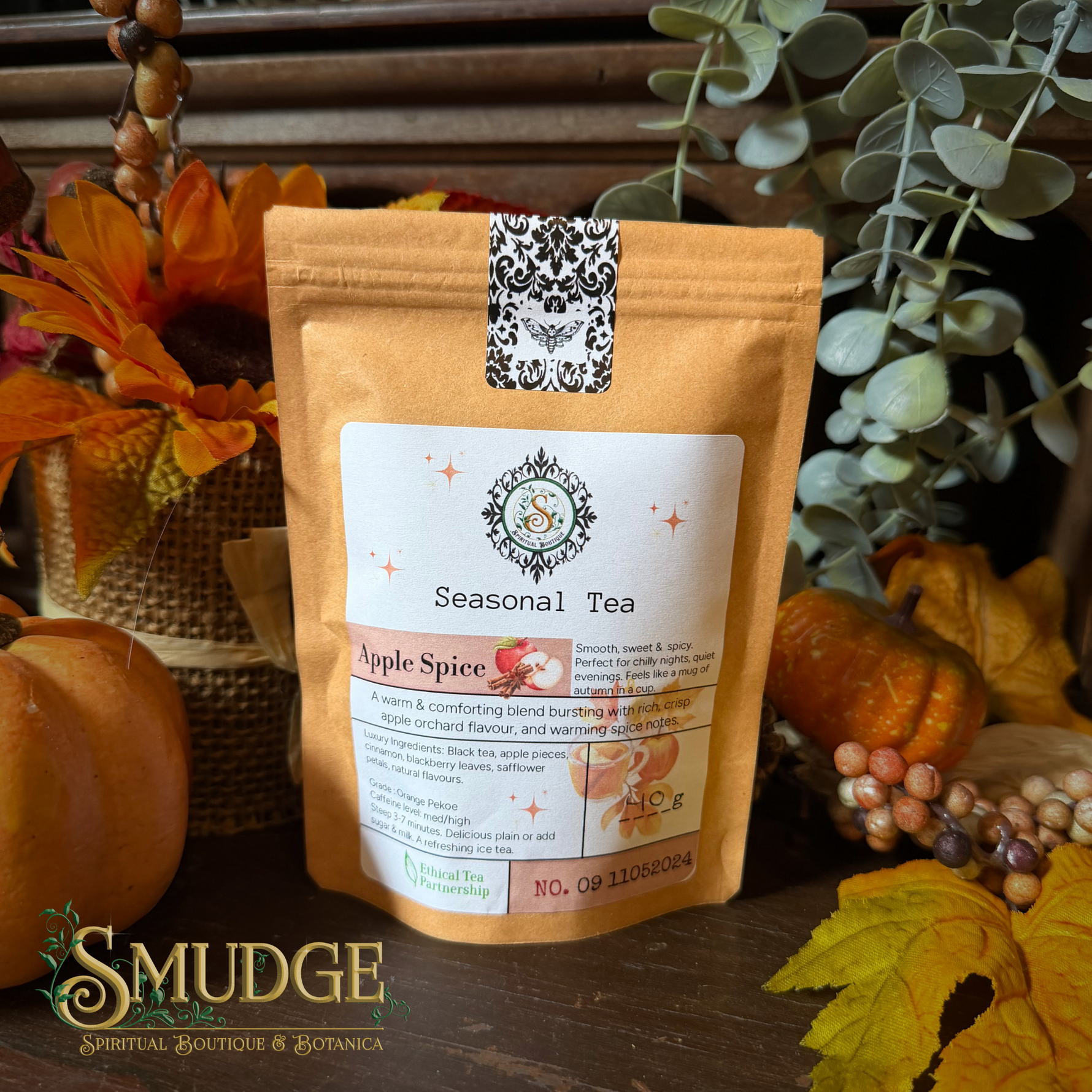 A stand-up pouch of Seasonal Tea by Smudge Spiritual Boutique & Botanica, featuring an Apple Spice flavor. The pouch is styled in an autumn setting with pumpkins, fall leaves, and eucalyptus on a wooden surface.