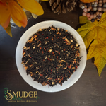 A top-down view of a white bowl filled with dark loose-leaf herbal tea mixed with dried citrus slices and small orange pieces, set on a dark wooden surface with autumn leaves, a pinecone, and berries around it. The brand logo 'SMUDGE SPIRITUAL BOUTIQUE & BOTANICA' is visible in the bottom-left corner.
