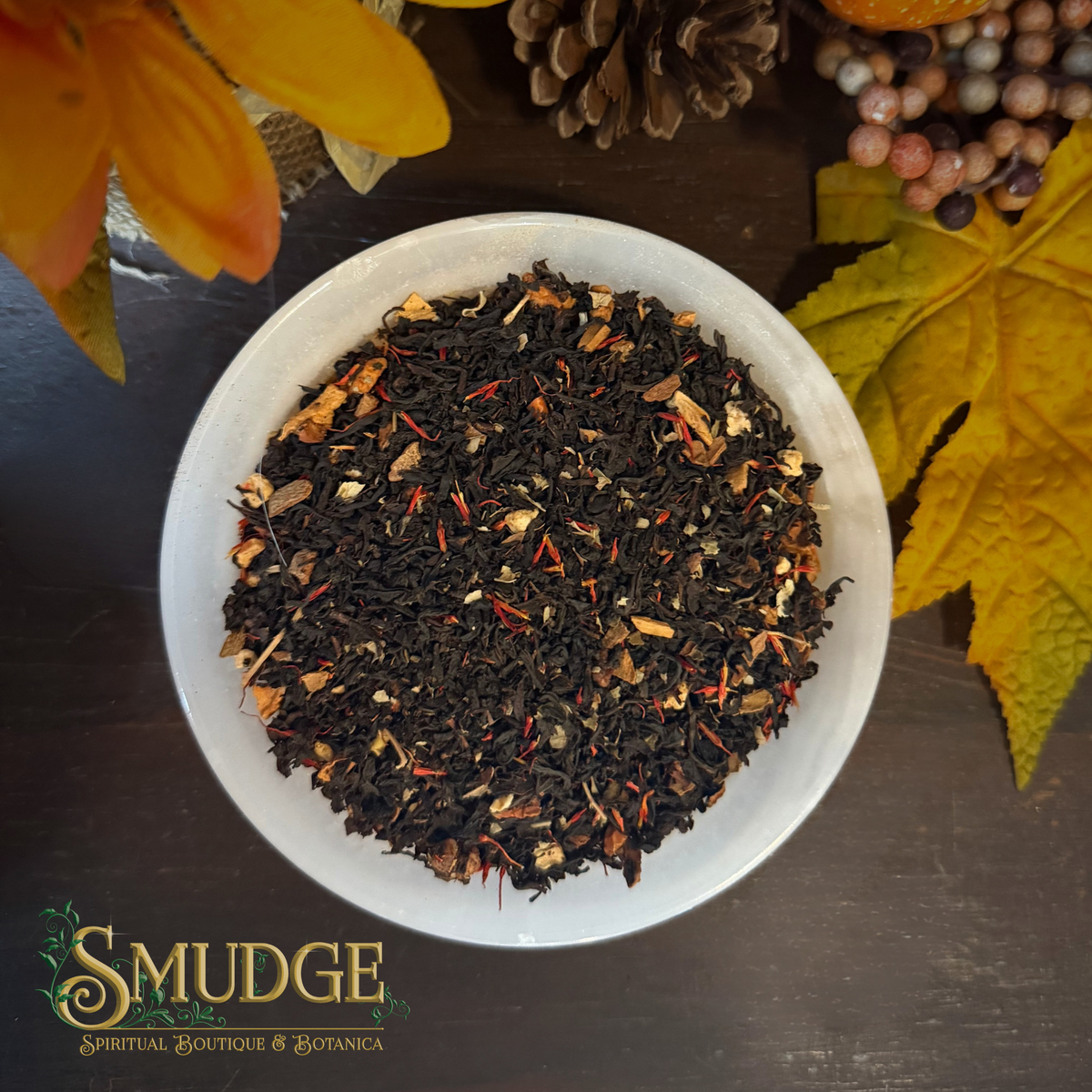 A top-down view of a white bowl filled with dark loose-leaf herbal tea mixed with dried citrus slices and small orange pieces, set on a dark wooden surface with autumn leaves, a pinecone, and berries around it. The brand logo 'SMUDGE SPIRITUAL BOUTIQUE & BOTANICA' is visible in the bottom-left corner.