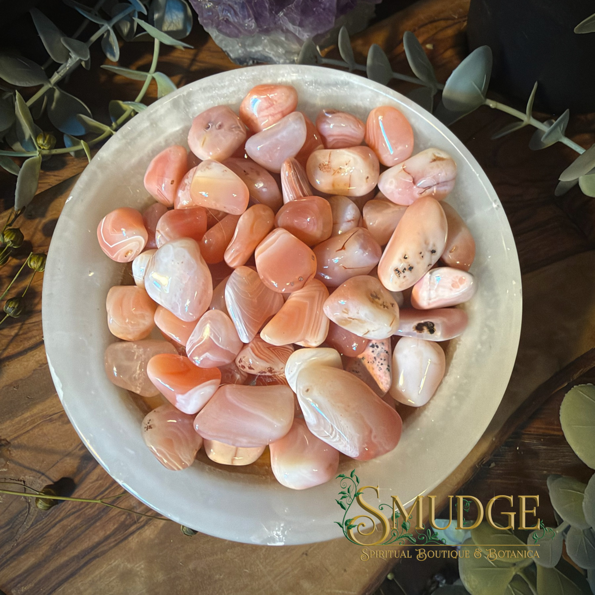 A bowl filled with polished peach, pink, and white agate tumbled stones, styled on a dark wooden surface with green eucalyptus leaves and a Smudge Spiritual Boutique logo in the corner.
