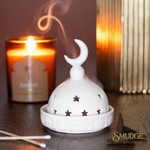 A white ceramic incense burner with a crescent moon and star cutouts, shown with incense sticks and a lit candle in a warm, atmospheric setting. The brand logo 'SMUDGE' is visible in the bottom right corner.