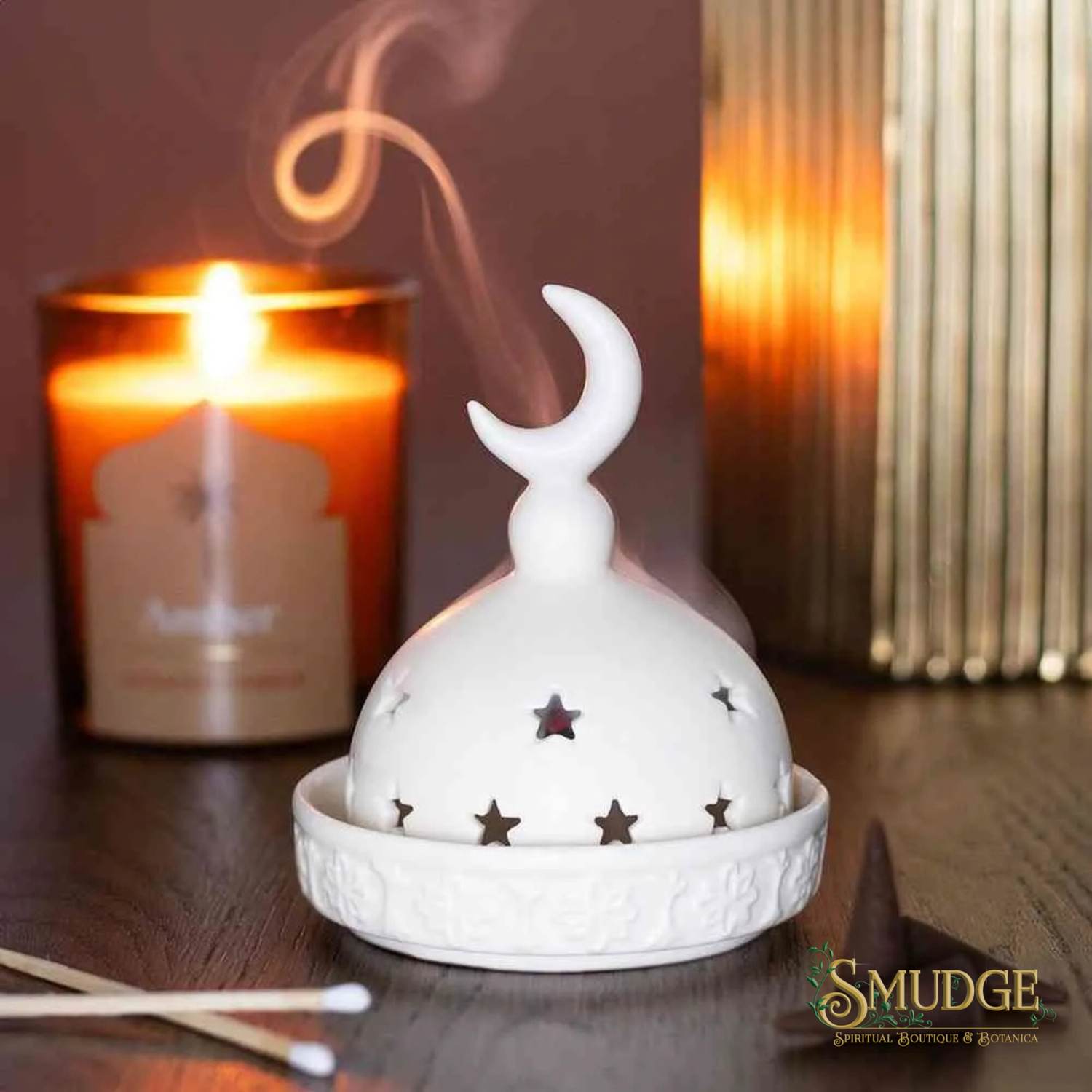 A white ceramic incense burner with a crescent moon and star cutouts, shown with incense sticks and a lit candle in a warm, atmospheric setting. The brand logo 'SMUDGE' is visible in the bottom right corner.