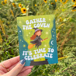 Hand holding a card with 'Gather the Coven, It's Time to Celebrate' text against a sunflower field background.