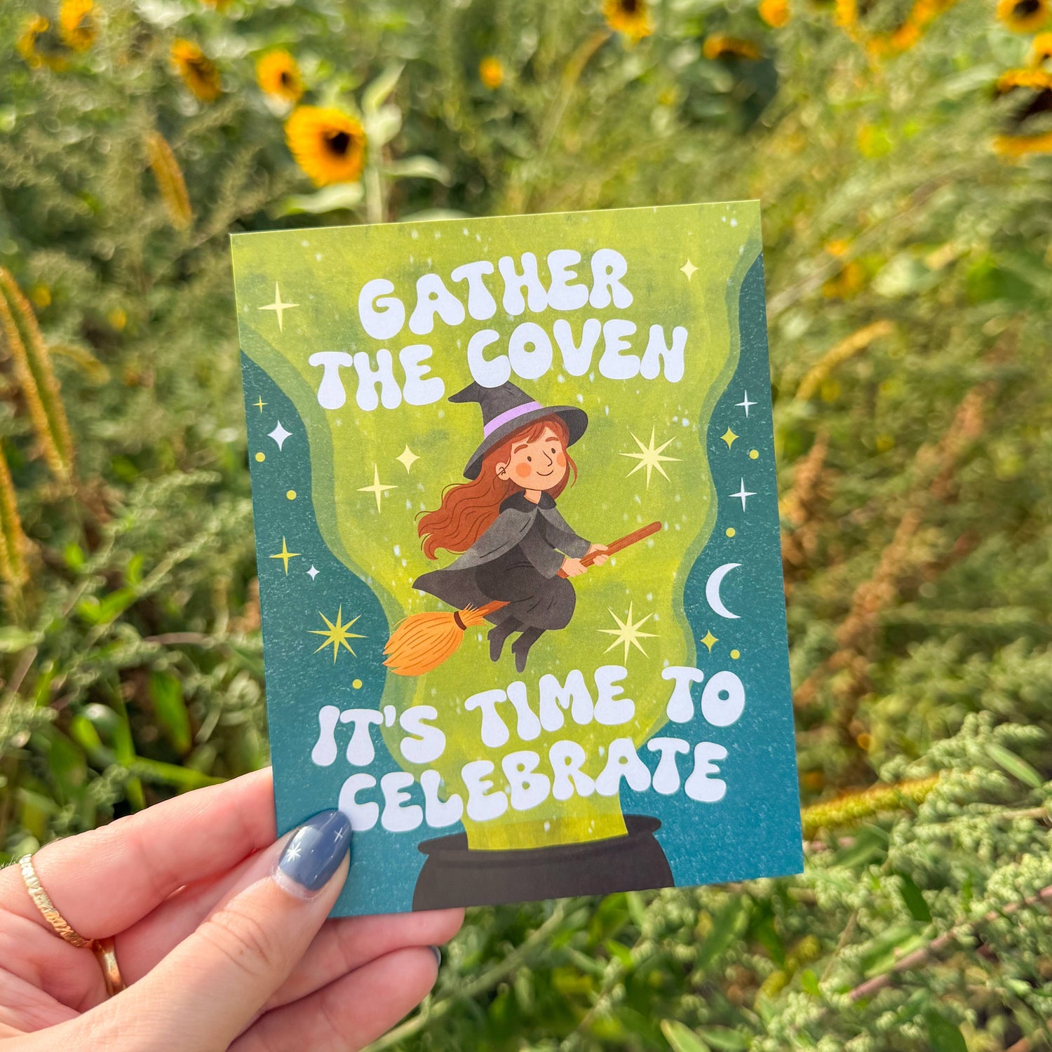 Hand holding a card with 'Gather the Coven, It's Time to Celebrate' text against a sunflower field background.