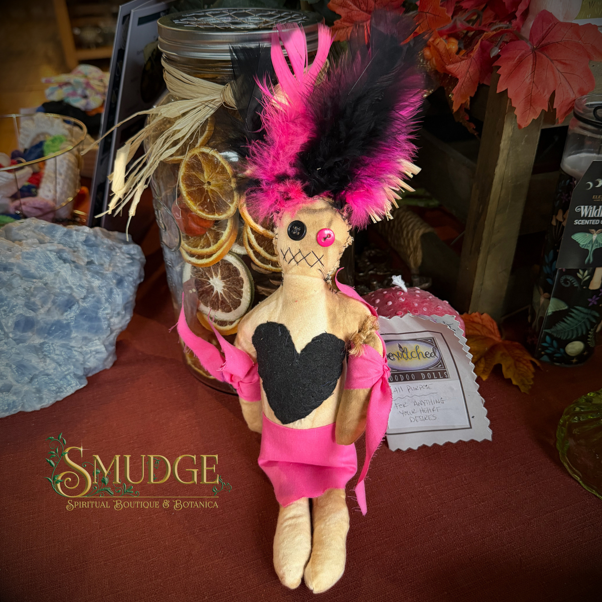A handmade rag doll with a black and pink feathered headdress, a pink dress, and a black heart on its chest, displayed on a table at a craft fair. A 'Smudge Spiritual Boutique & Botanica' logo is visible in the bottom left corner.