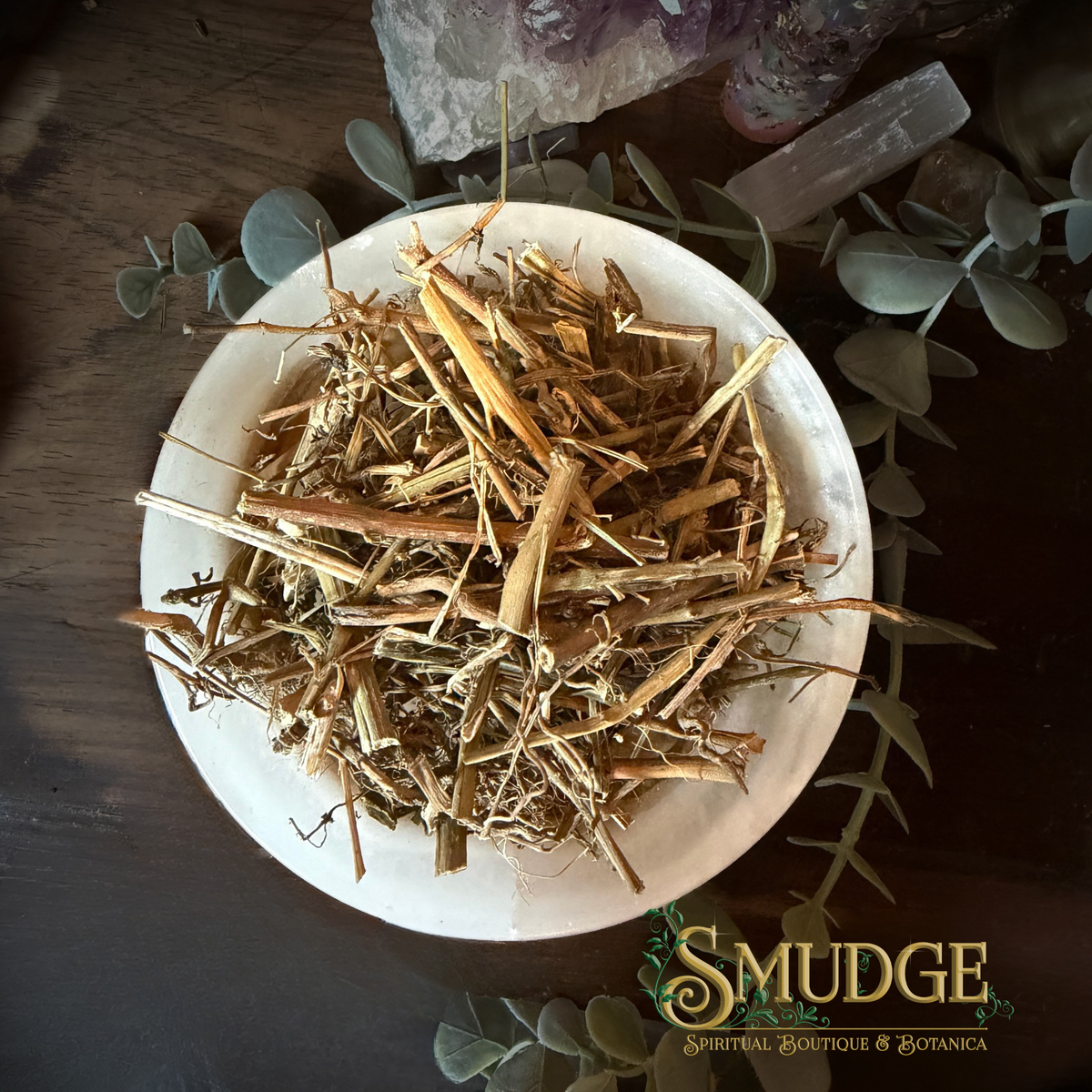 A top-down view of dried botanical roots and stems piled in a white ceramic dish, surrounded by eucalyptus leaves and crystals on a dark wooden surface. The brand logo 'SMUDGE SPIRIT BOUTIQUE & BOTANICA' is visible in the bottom right corner.