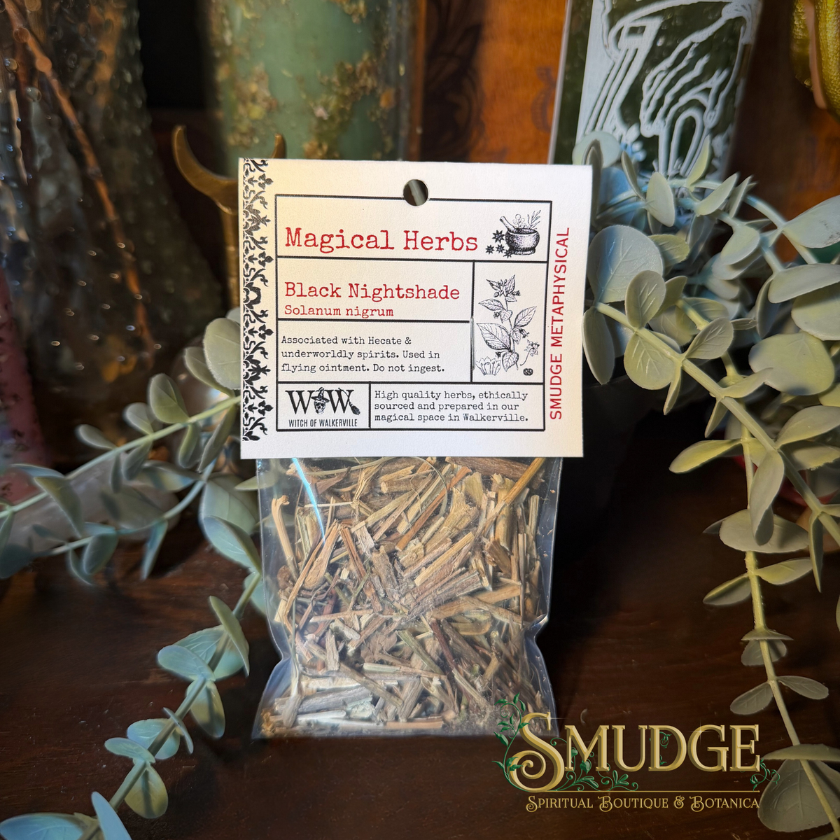 A clear plastic bag filled with dried botanical herbs, sealed with a white paper label reading 'Magical Herbs' and 'Black Nightshade', photographed on a dark wooden surface with eucalyptus leaves and decorative objects in the background.