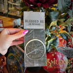 A hand with pink nail polish holds a packaged circular air freshener. The clear plastic packaging shows a round white disc with black pentagram and moon illustrations, along with a gray header card reading 'BLESSED BE AIR FRESHENER' and 'VANILLA SCENTED AIR FRESHENER.
