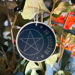 A round dark blue hanging ornament or tag with a white pentagram and the words 'BLESSED BE' in white, hanging among green leaves and branches.