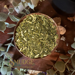 A top-down view of a brown ceramic bowl filled with dried green herbal chopped grass, surrounded by eucalyptus leaves and stones on a dark wooden surface. The brand logo 'SMUDGE' is visible in the bottom-left corner.