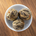 Three intricately wrapped tea balls in a white bowl with more tea balls around.