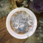 A top-down view of a white ceramic bowl filled with polished clear tumbled stones, set on a wooden surface with decorative greenery and an amethyst cluster. The SMUDGE Spiritual Boutique & Botanicals logo is visible in the bottom right corner.
